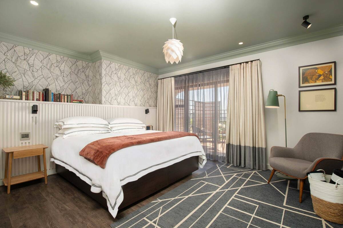 Un dormitorio con una cama grande, una mesita de noche, una silla, una lámpara de pie y una ventana con cortinas.