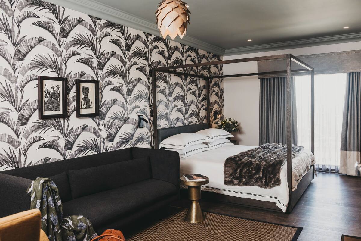 Un dormitorio con una cama con dosel, un sofá oscuro, una mesita de noche pequeña y un ventanal con cortinas.