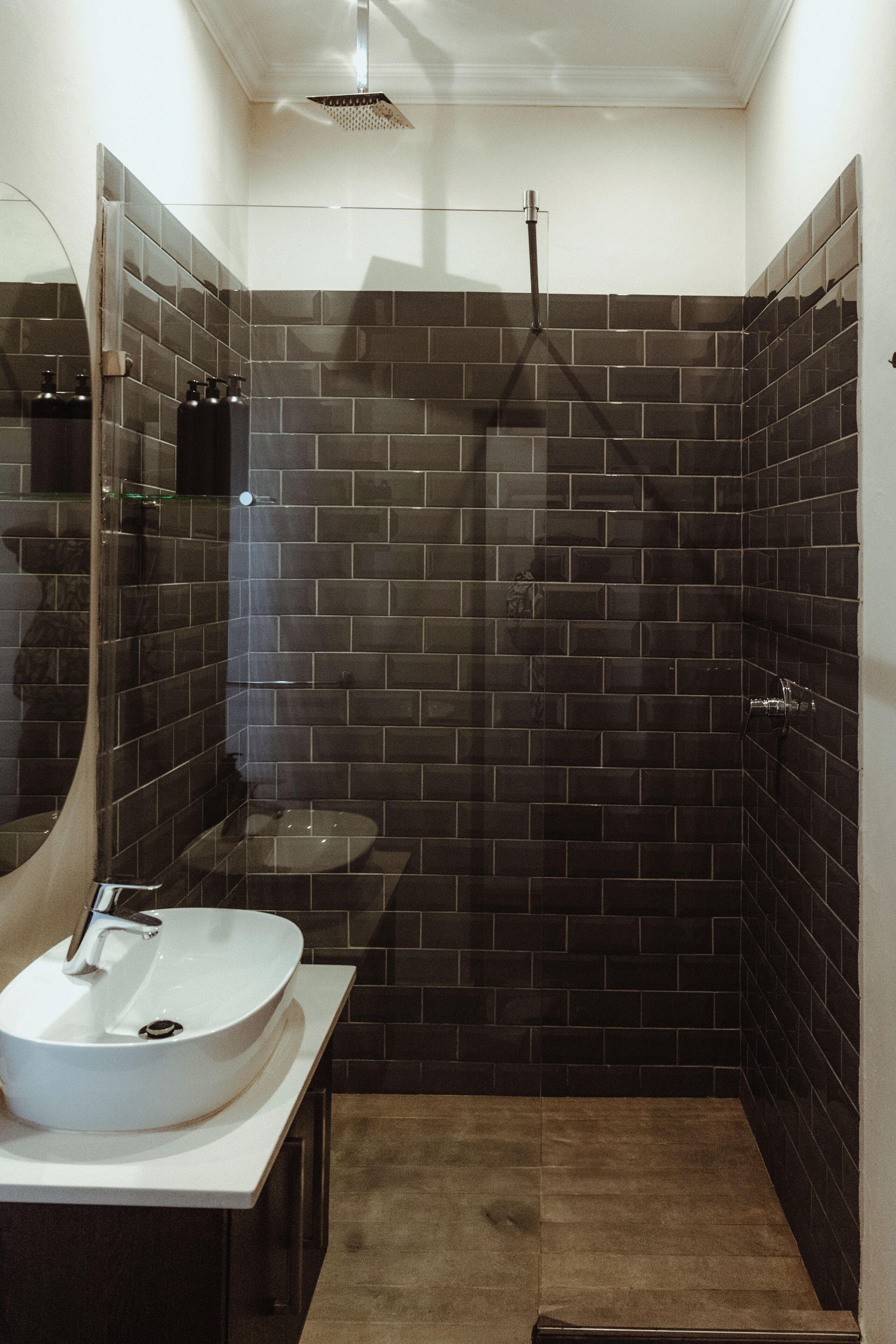 Un baño moderno con un lavamanos blanco, paredes cubiertas de azulejos oscuros y un box de ducha de vidrio.