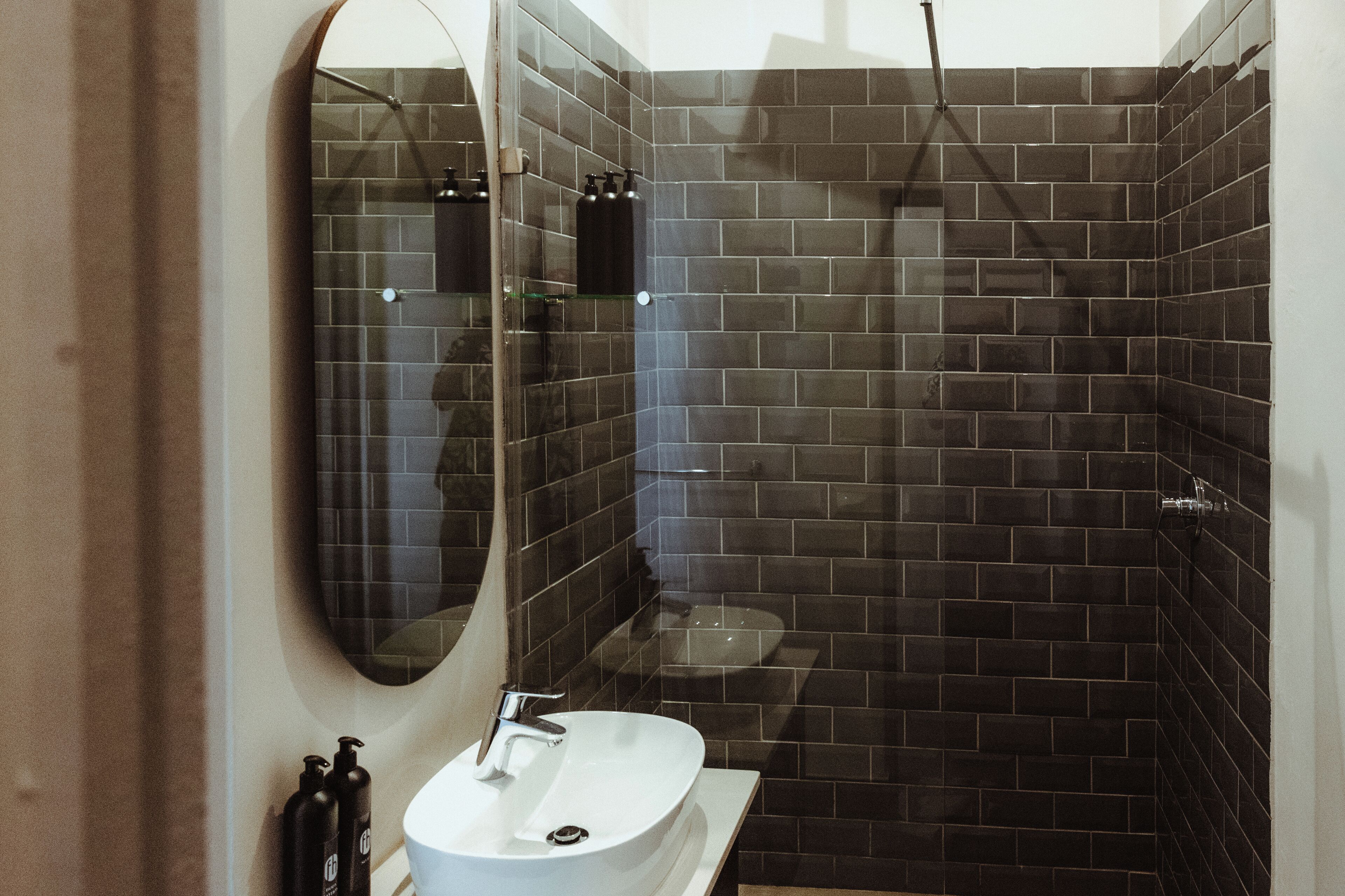 Une salle de bain moderne avec un lavabo blanc, un grand miroir et des murs recouverts de carreaux foncés.