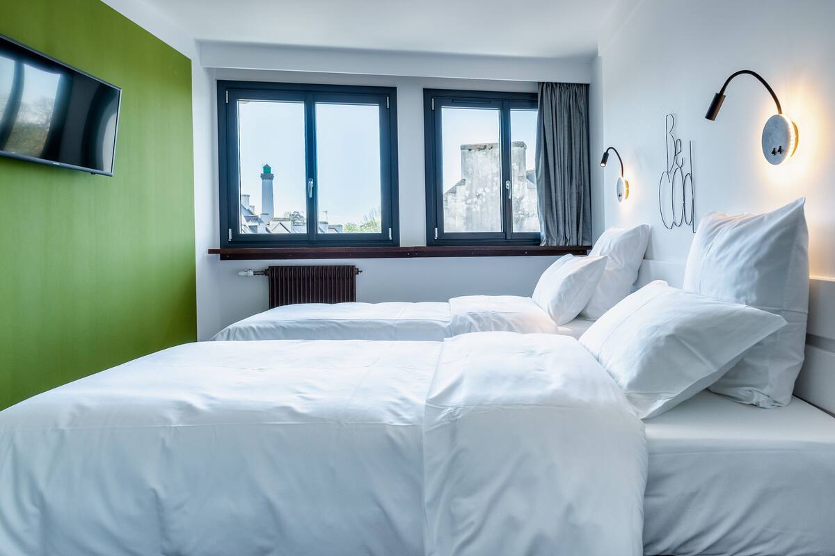 Habitación de hotel moderna con una cama grande, dos ventanas, un televisor de pantalla plana y vistas a un paisaje urbano.