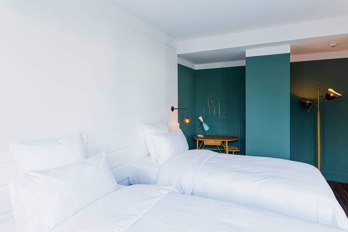 Una cama bien hecha con ropa blanca en una habitación con paredes verde azulado y una pequeña mesa de madera.