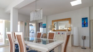 A modern dining area with a glass-top table, white chairs with blue cushions, a chandelier, and a large mirror on the wall. - Casa Monsoria - sea views, private pool, walking distance to beach & town centre (Carvoeiro)