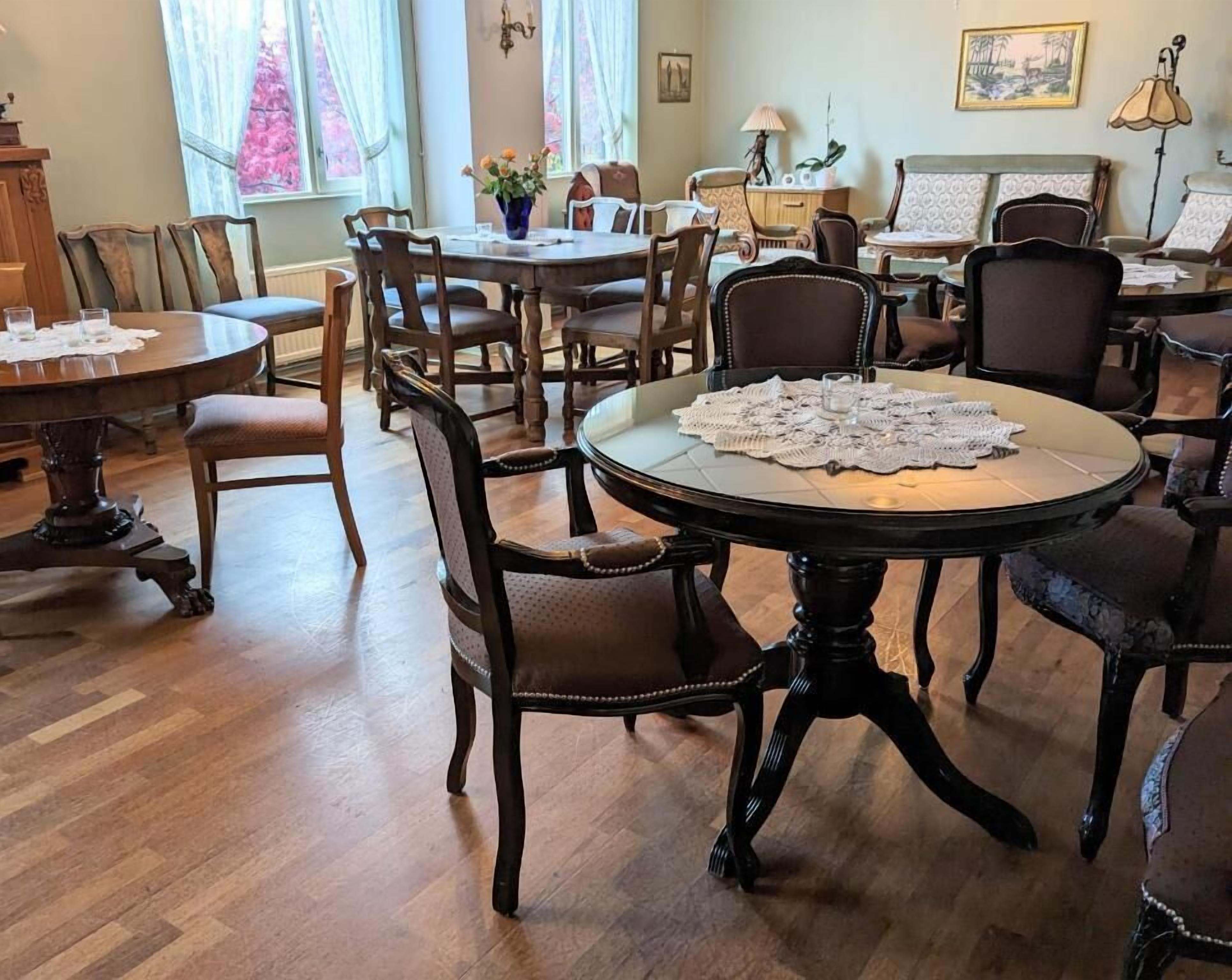 A dining area with multiple wooden tables and chairs, a lace table runner on one table, and a window with curtains.