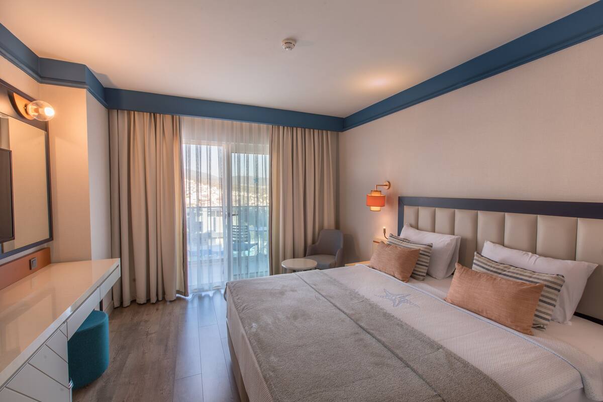 A modern hotel room with a large bed, a desk, and a chair. There is a balcony with curtains and a view of the city.
