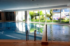 Une piscine intérieure aux parois en verre transparent, dotée d’un sol carrelé et offrant une vue sur un espace extérieur avec piscine, de la verdure et une statue.