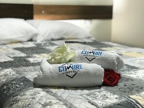Un lit avec une couvre-lit matelassé, deux serviettes portant la mention « CUMBRE HOTEL », et une rose rouge.