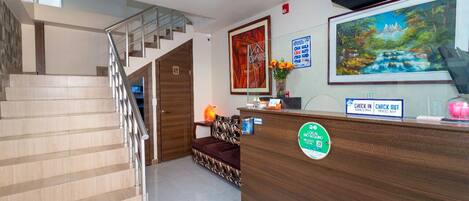 A reception area with a wooden desk, a check-in/check-out counter, a staircase, and a framed painting on the wall.