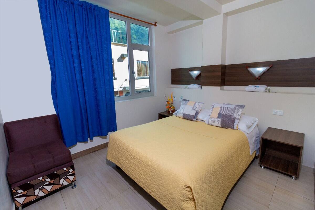 A bedroom with a bed, a nightstand, a chair, a window with curtains, and a wall-mounted shelf.