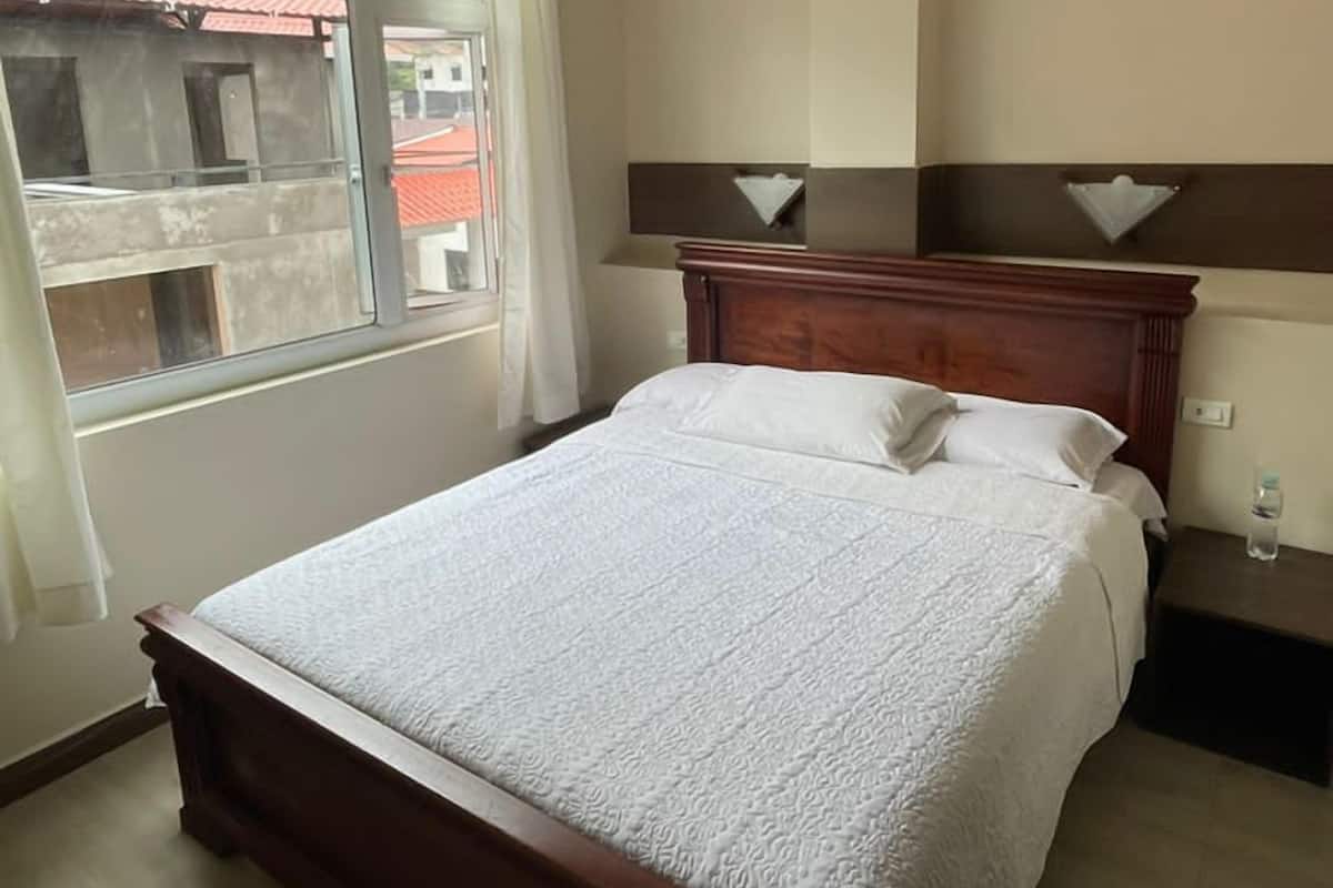 A bedroom with a large bed, a nightstand, a bedside table, a window with curtains, and a ceiling light.