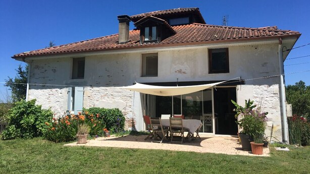 Una casa a due piani con tetto in tegole, un portico coperto con tavolo e sedie, e piante in vaso.