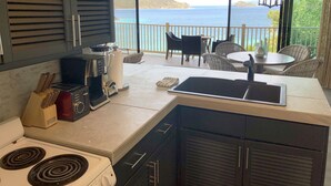Una cocina con estufa, cafetera y tostadora. Hay un comedor con sillas y una mesa, y vista al océano.