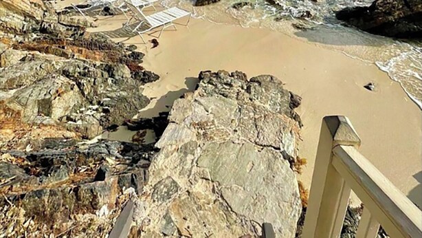 Una playa con agua turquesa cristalina, costa rocosa y un paseo de madera que lleva a sillas de playa.