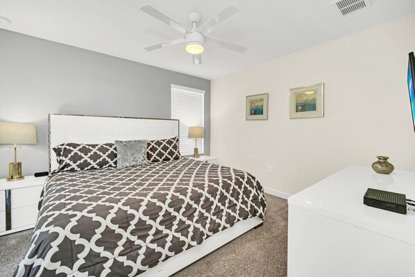 A bedroom with a bed, a nightstand, a desk, a ceiling fan, and two wall-mounted paintings.