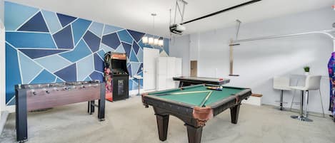 Una sala recreativa con mesa de billar, mesa de futbolín y una máquina arcade colorida.