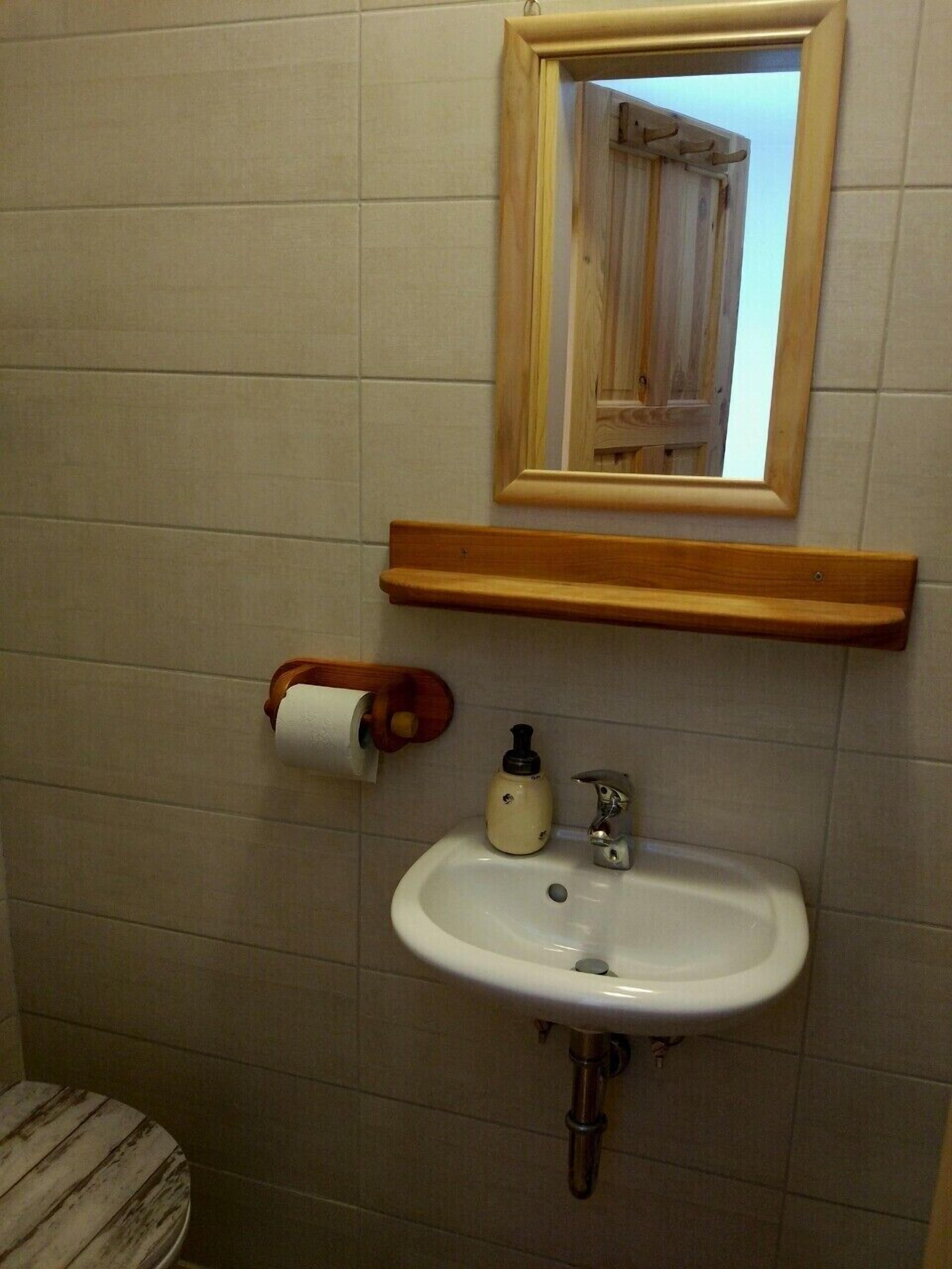 Uma casa de banho com uma pia branca, um espelho, uma sanita e uma prateleira de madeira.