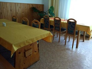 A dining area with a yellow tablecloth-covered table, wooden chairs with patterned cushions, and a large plant in the background.