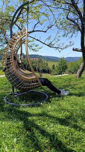 A person relaxing in a large, curved wicker chair with a circular base, set on a grassy area with trees and a distant view of hills.