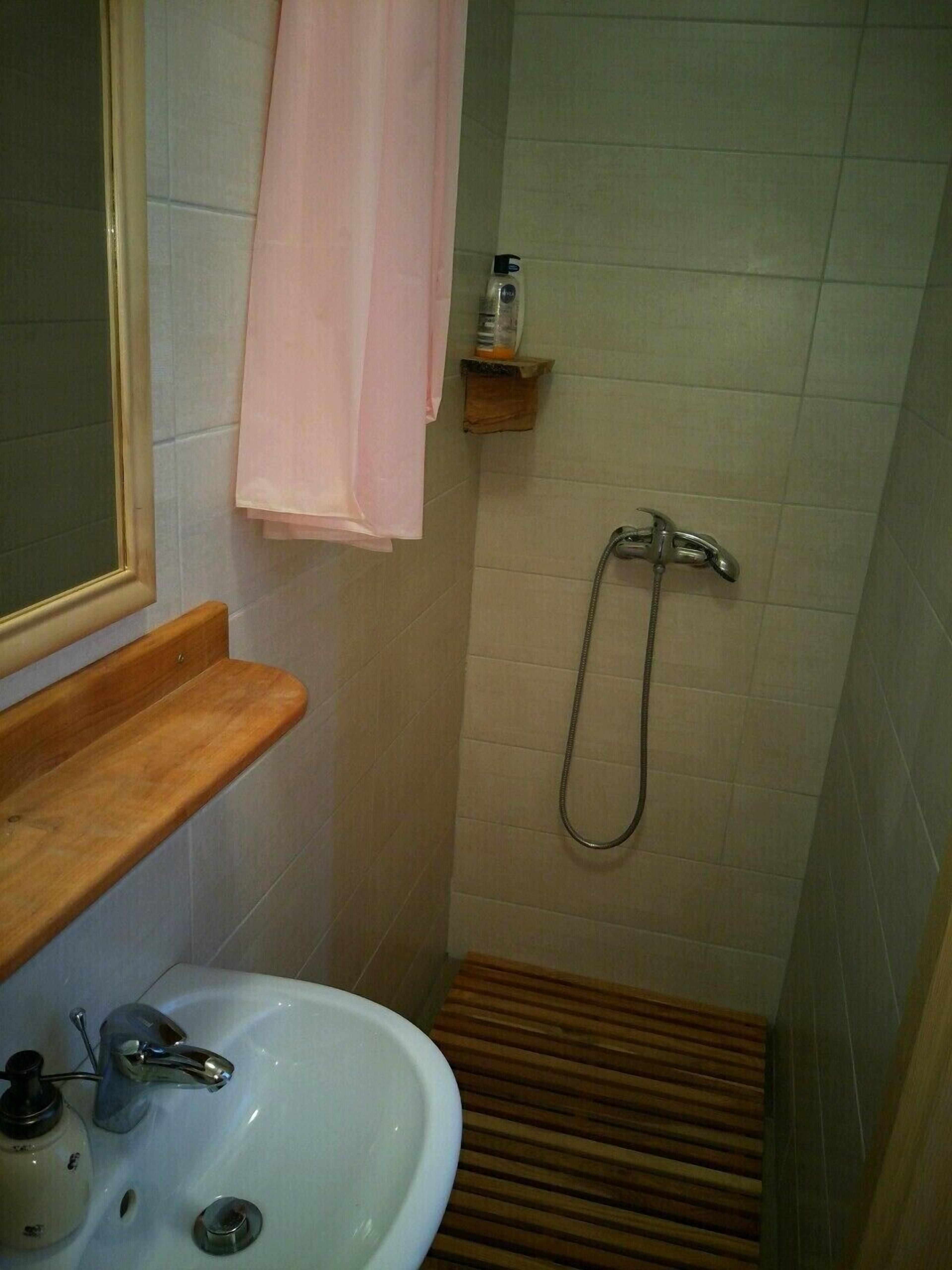 A bathroom with a sink, mirror, shower, and wooden shelf.