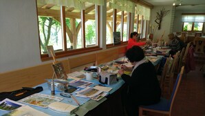 A room with a long table set up for an art class, featuring various art supplies and papers.