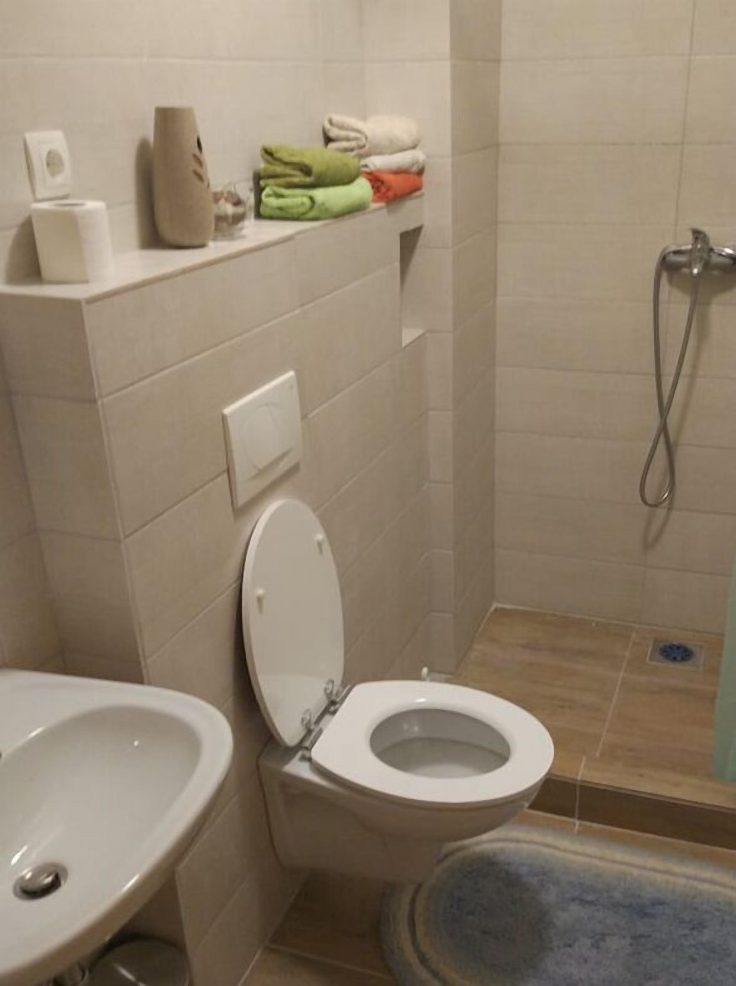 A bathroom with a toilet, sink, and shower. There are towels and toiletries on a shelf.