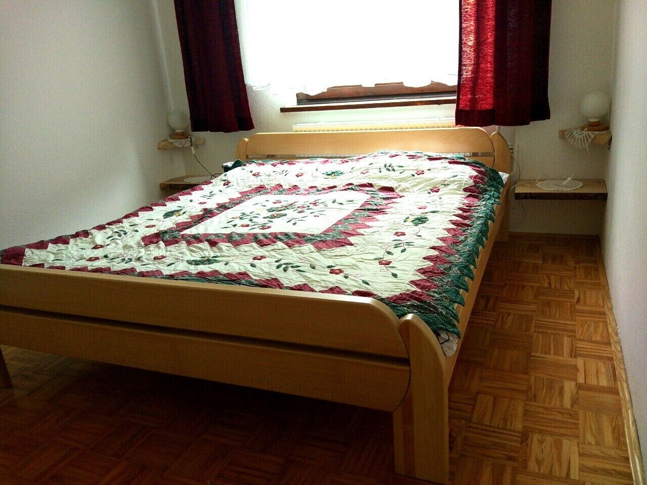 Una habitación pequeña con cama individual, estructura de cama de madera, colcha estampada con motivos florales y ventana con cortinas rojas.