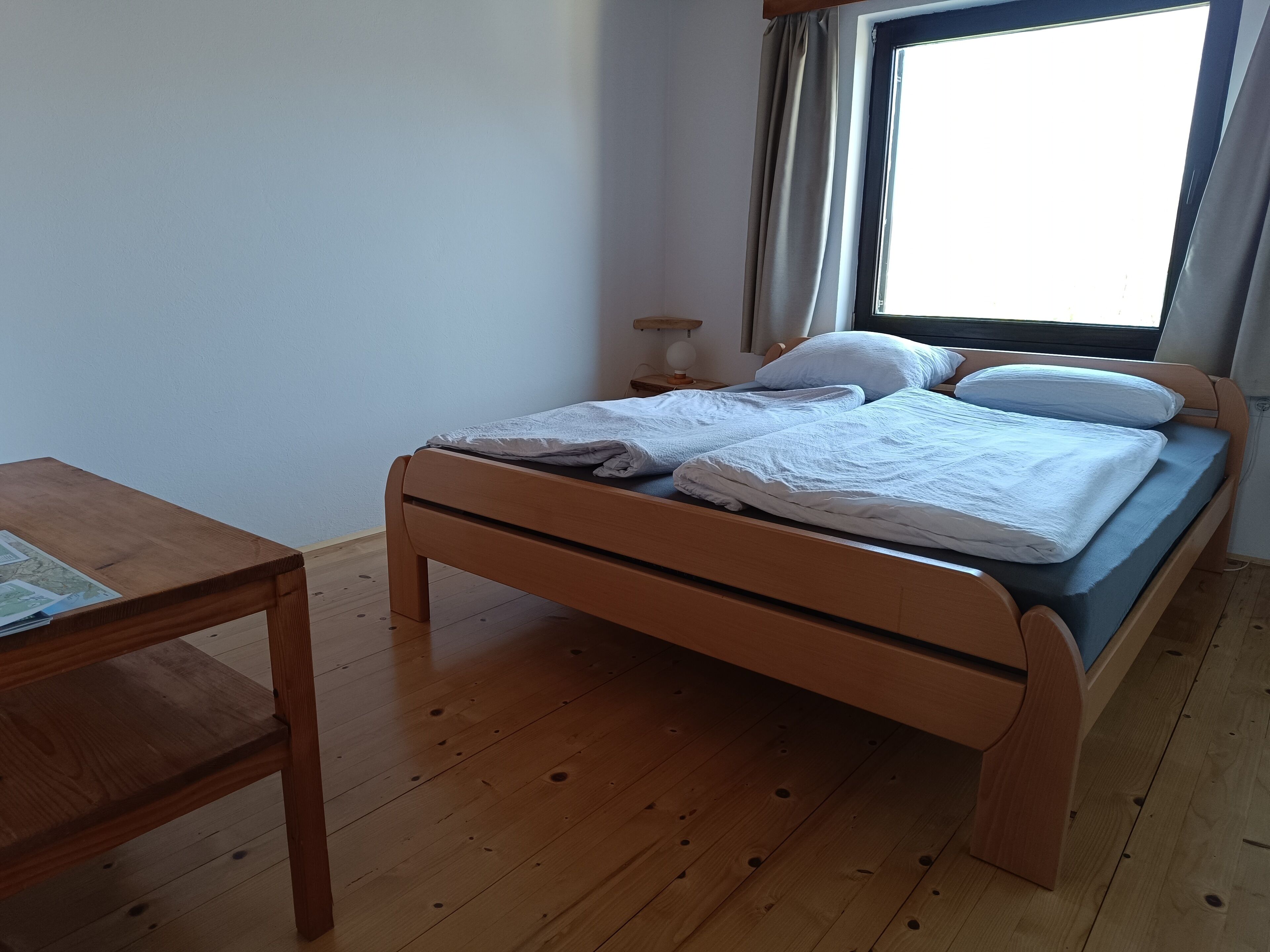 A single bed with a wooden frame and a blue mattress, a wooden nightstand with a lamp, and a small table with a map.