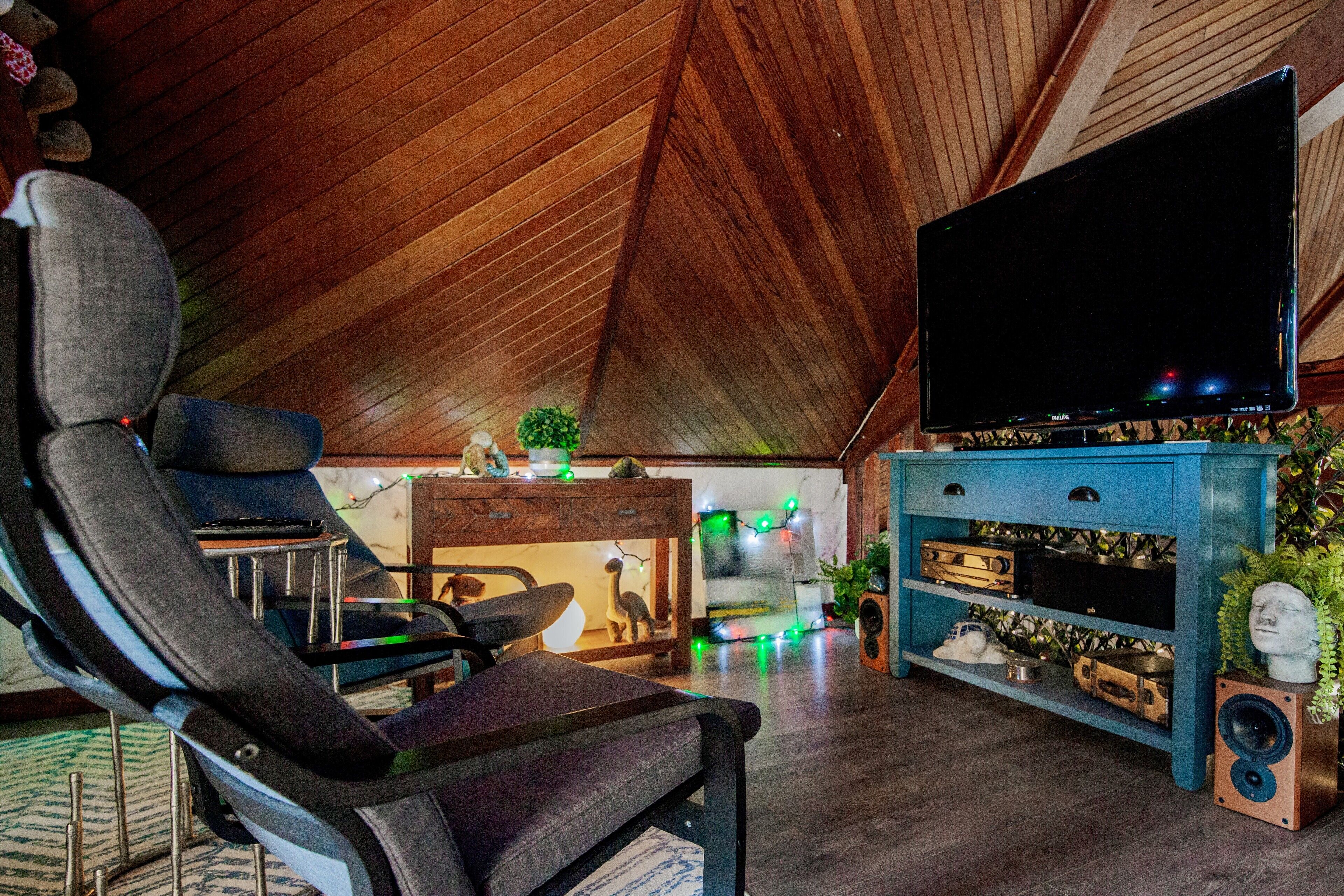 Una sala de estar acogedora con techo alto, un televisor de pantalla plana grande, un sillón cómodo, un mueble de entretenimiento azul y una pequeña planta.