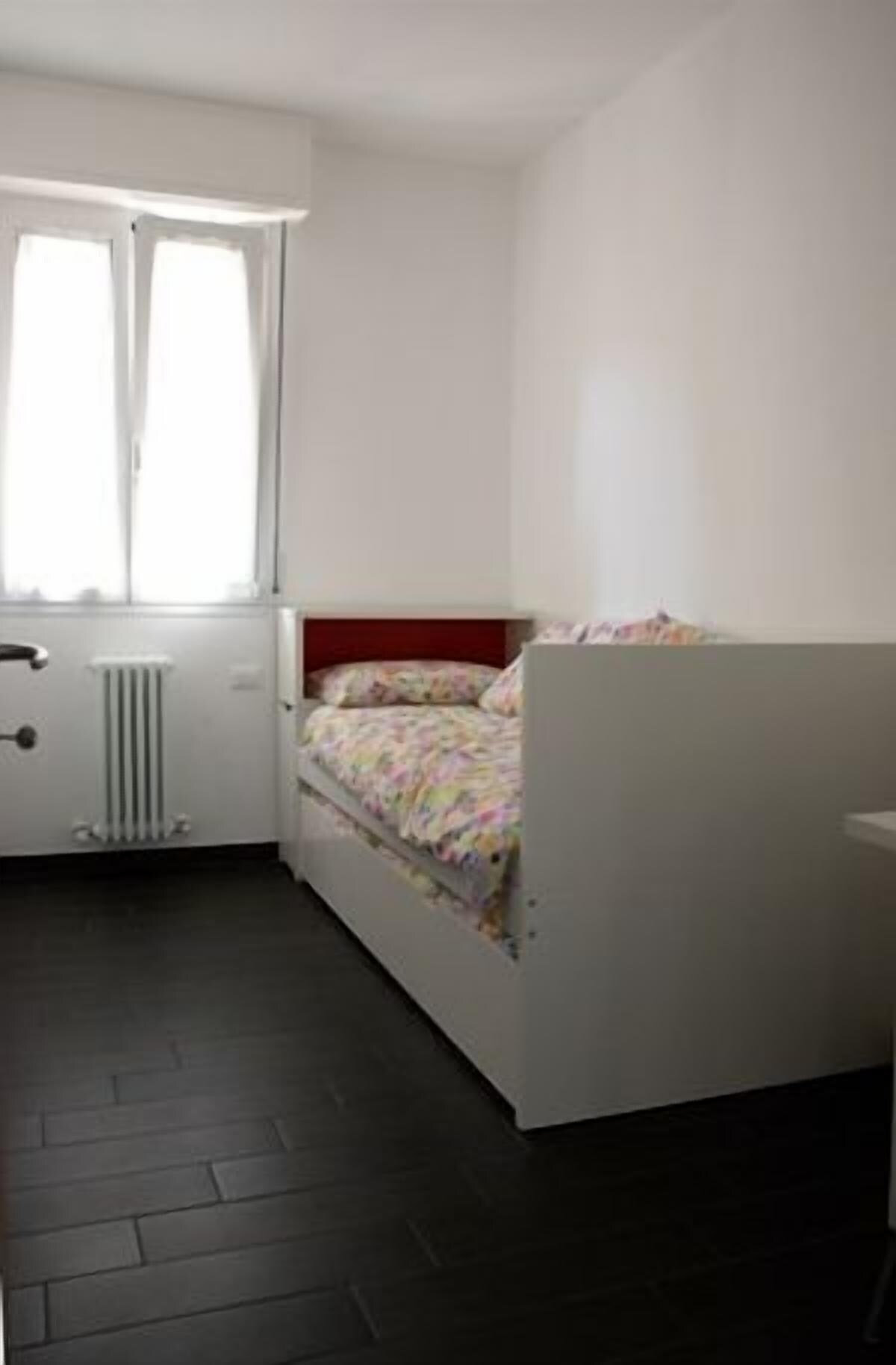 A small, single-bed room with a white headboard, a radiator below the window, and a dark wooden floor.