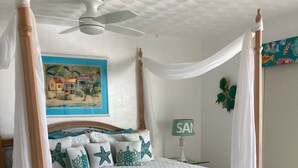 Una cama con dosel, un ventilador de techo, un cuadro de una playa y una mesita con superficie de vidrio.