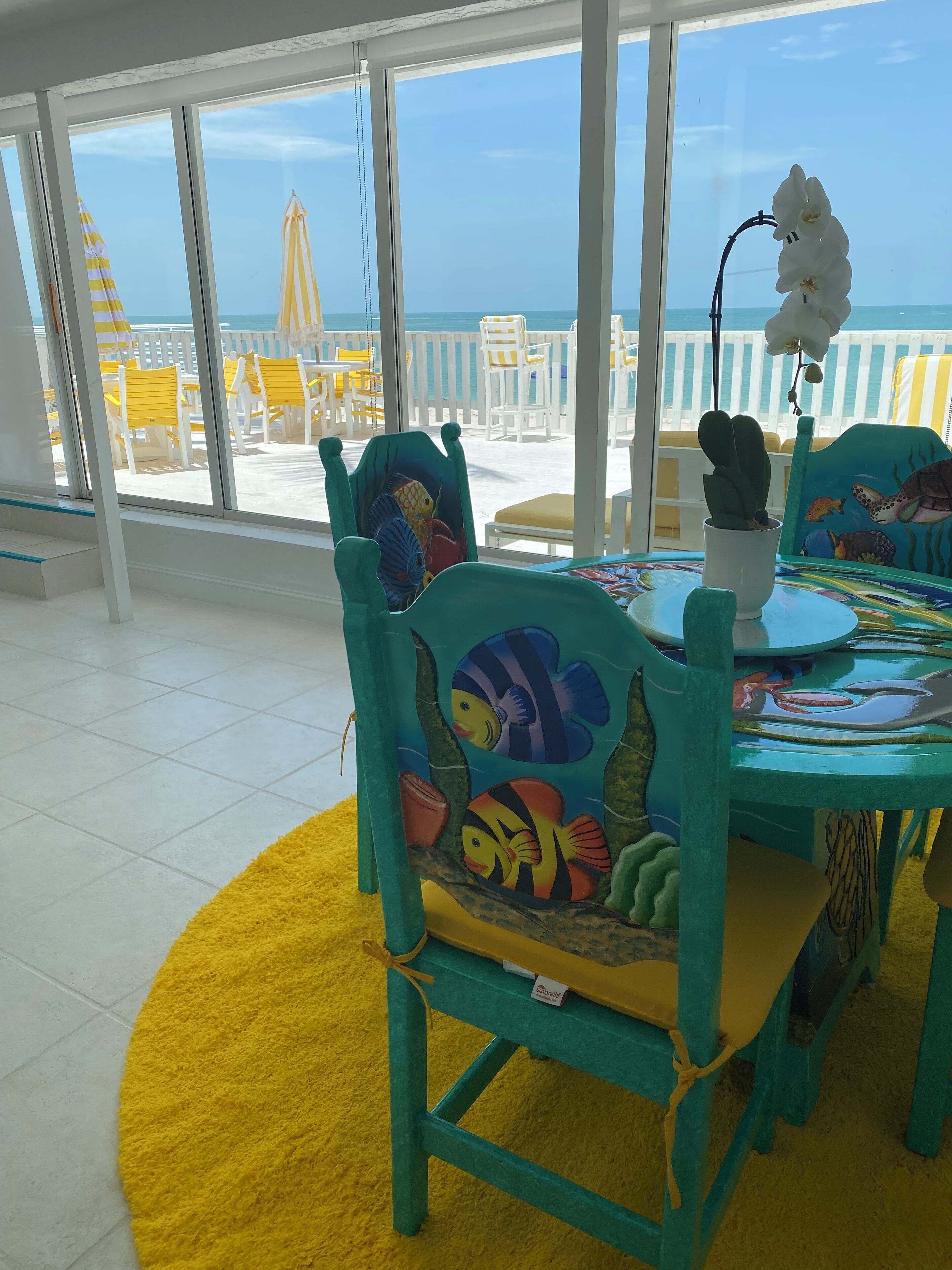 A dining area with a view of the beach, featuring a table with chairs decorated with fish designs, a yellow rug, and a potted orchid.