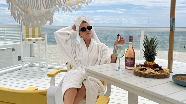 Una persona sentada a una mesa con una sombrilla de playa a rayas, una botella de vino y una bandeja de fruta.