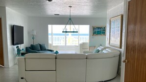 Un salón amplio con un sofá modular blanco grande, un ventilador de techo y vista al mar a través de amplios ventanales.