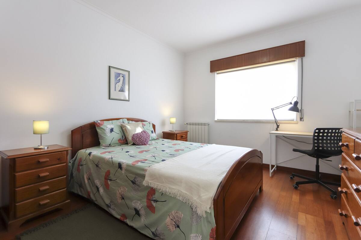A bedroom with a bed, a desk, a chair, a dresser, and a window with blinds.