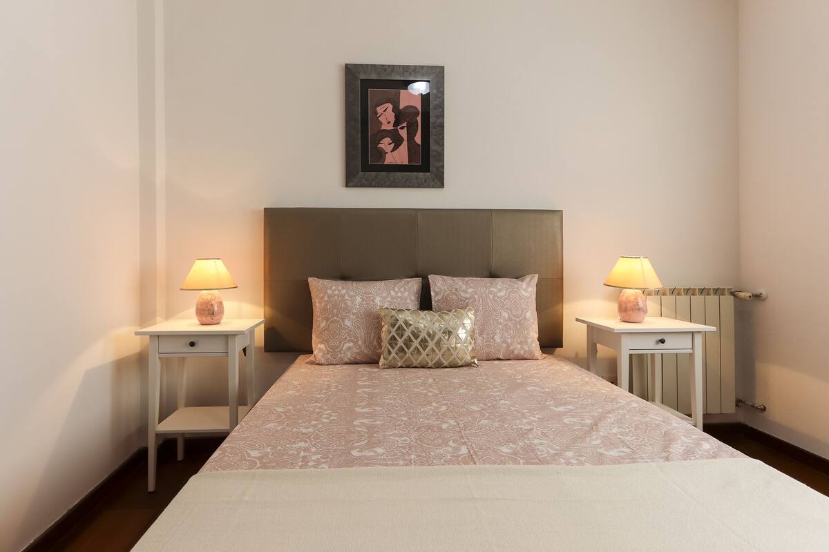 A neatly made bed with a patterned bedspread and two pillows, flanked by two bedside tables with lamps, in a simple room with a framed artwork above the bed.