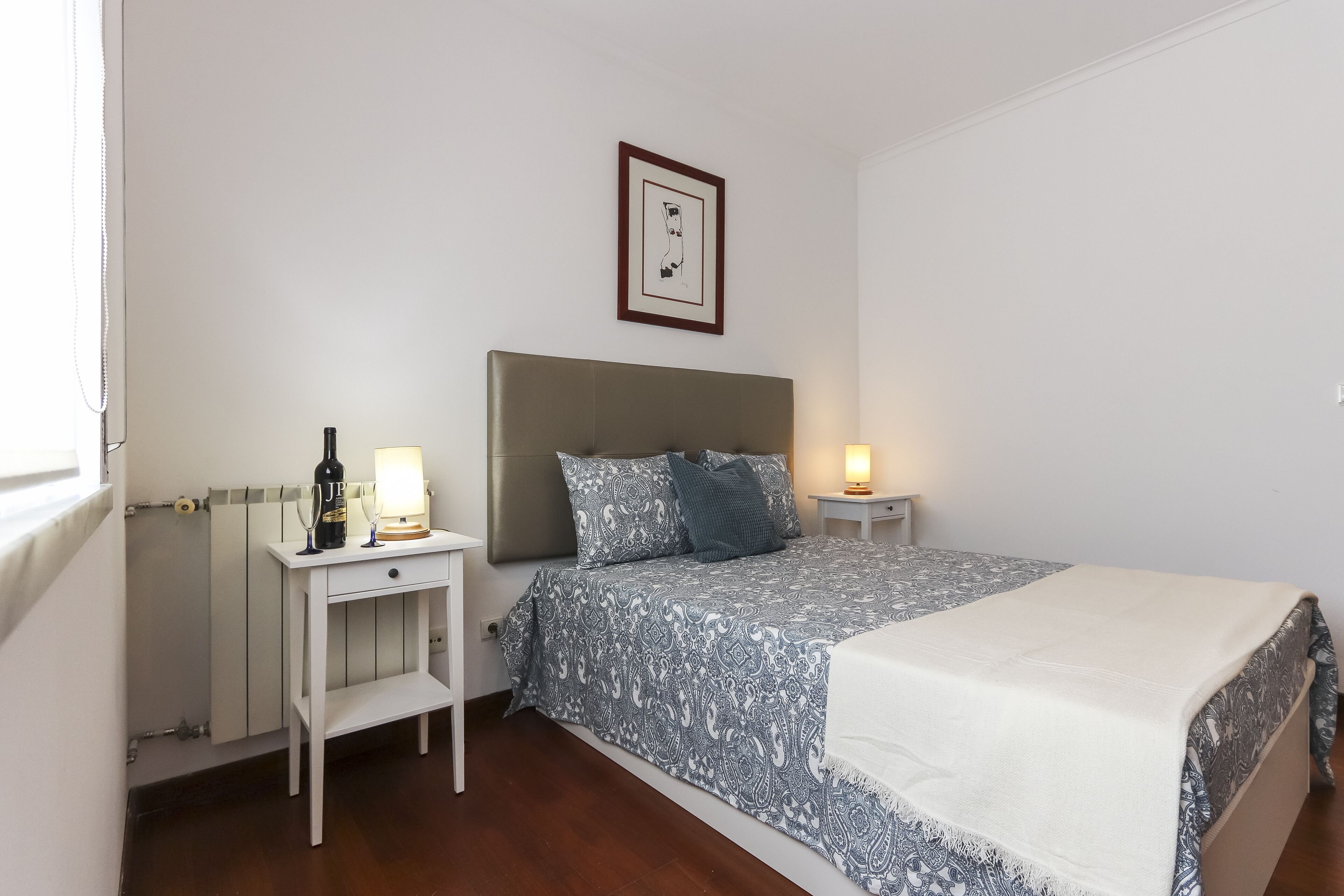 A bedroom with a bed, bedside tables, a nightstand, a framed picture on the wall, and a radiator.
