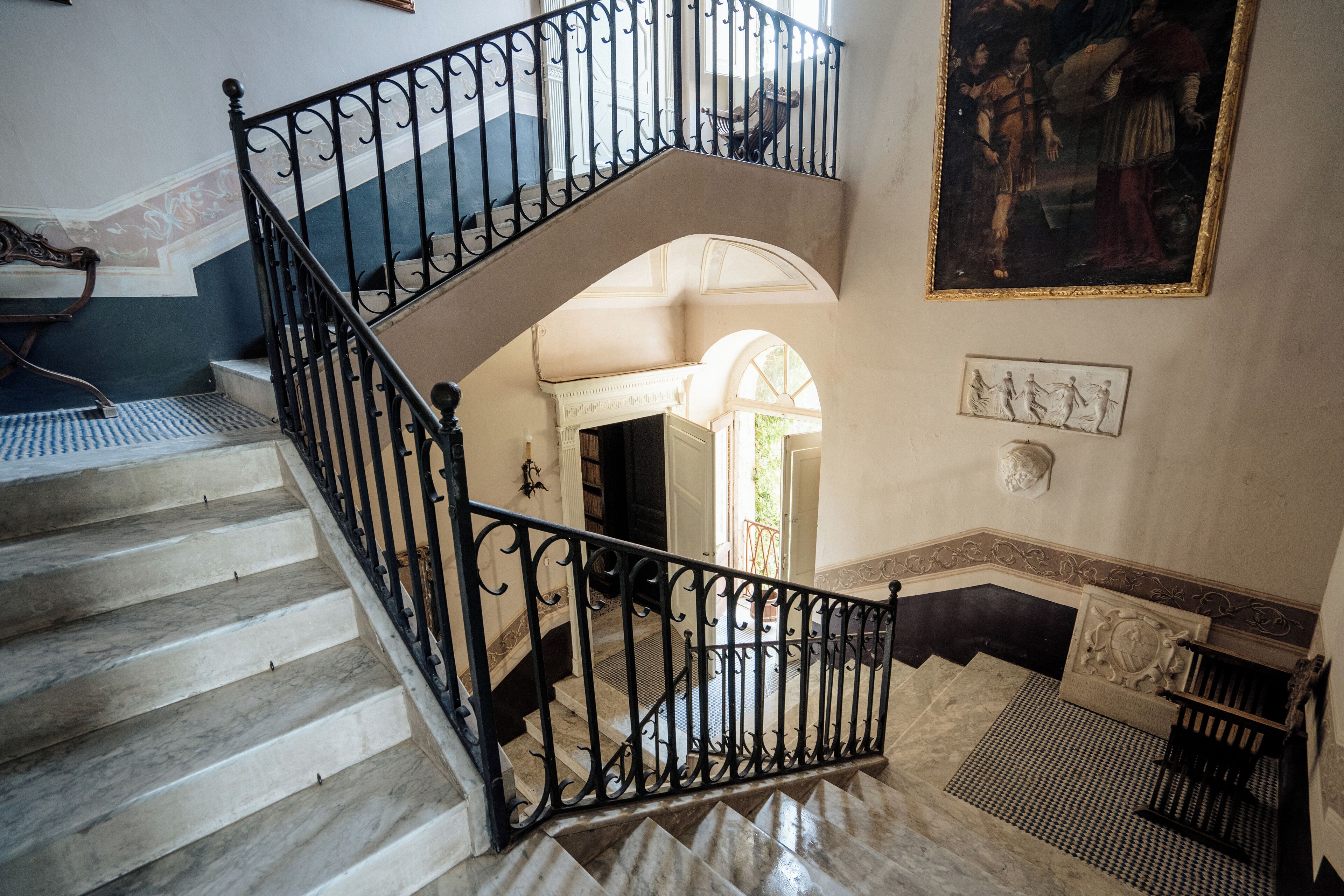 Una imponente escalera con barandales negros ornamentados, escalones de mármol y vistas a una habitación contigua con una puerta y una obra de arte enmarcada en la pared.