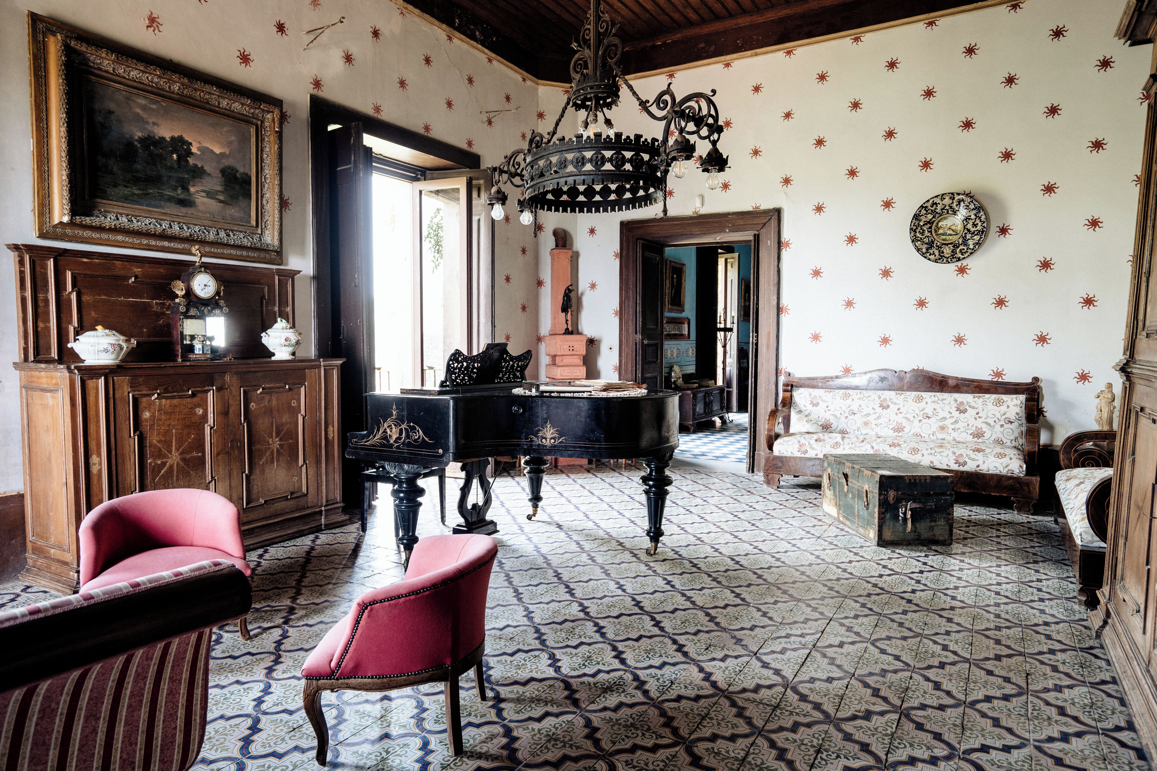 A room with a piano, a sofa, a red armchair, a wooden cabinet, and a chandelier.