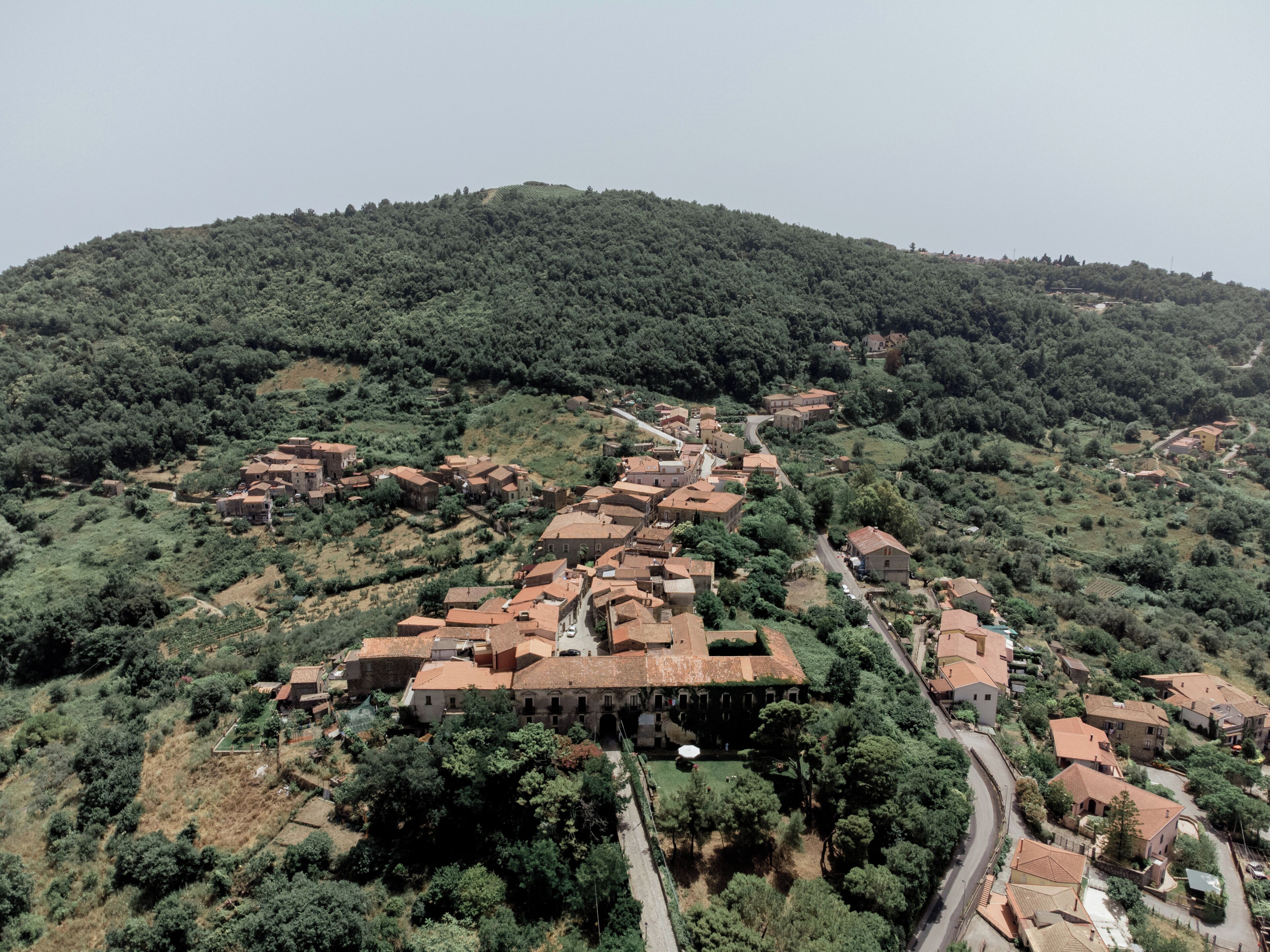 A village with terracotta-roofed buildings, narrow winding roads, and lush green hills in the background.