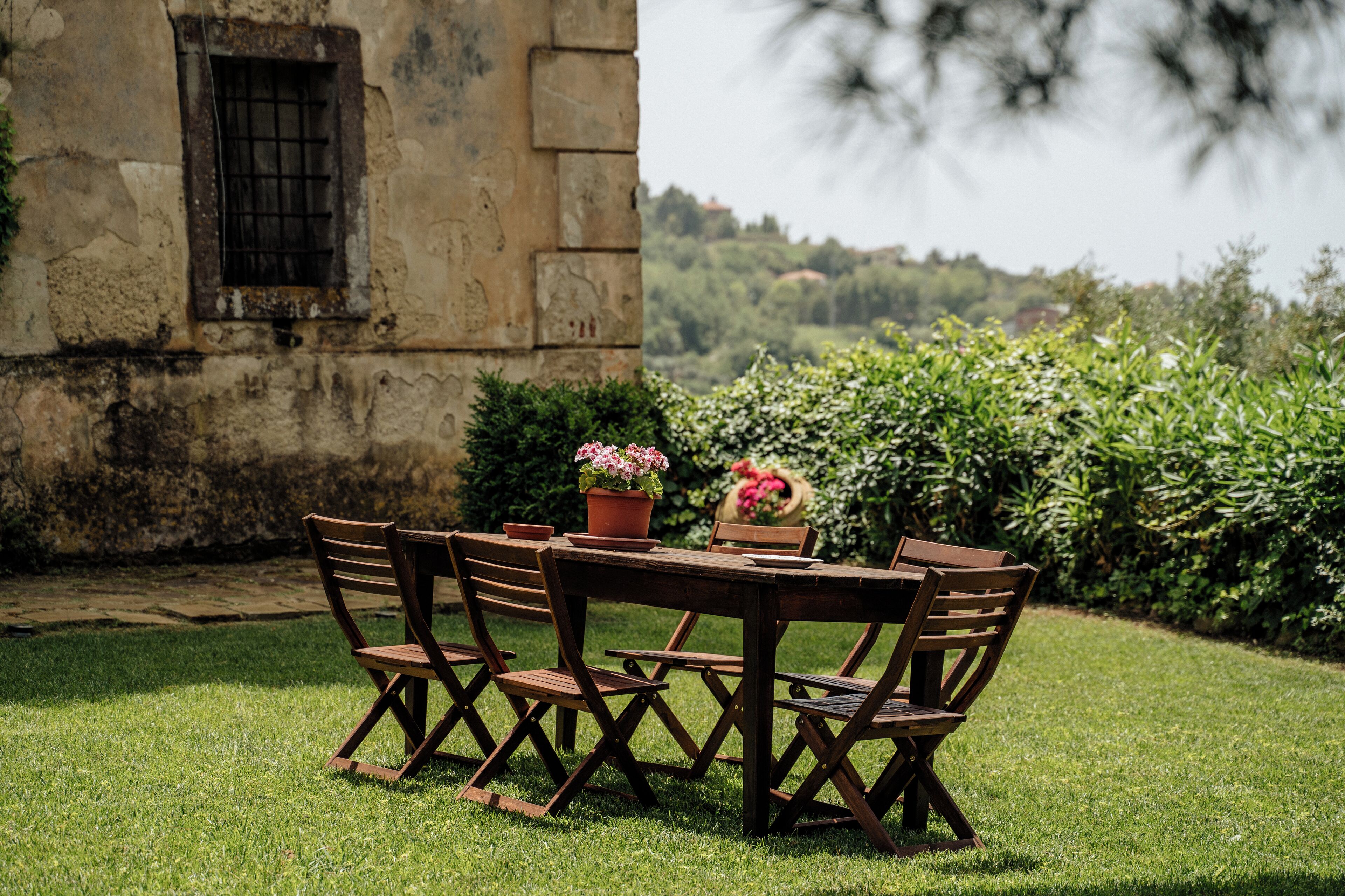 A wooden table and chairs set outdoors with a stone wall and greenery in the background.