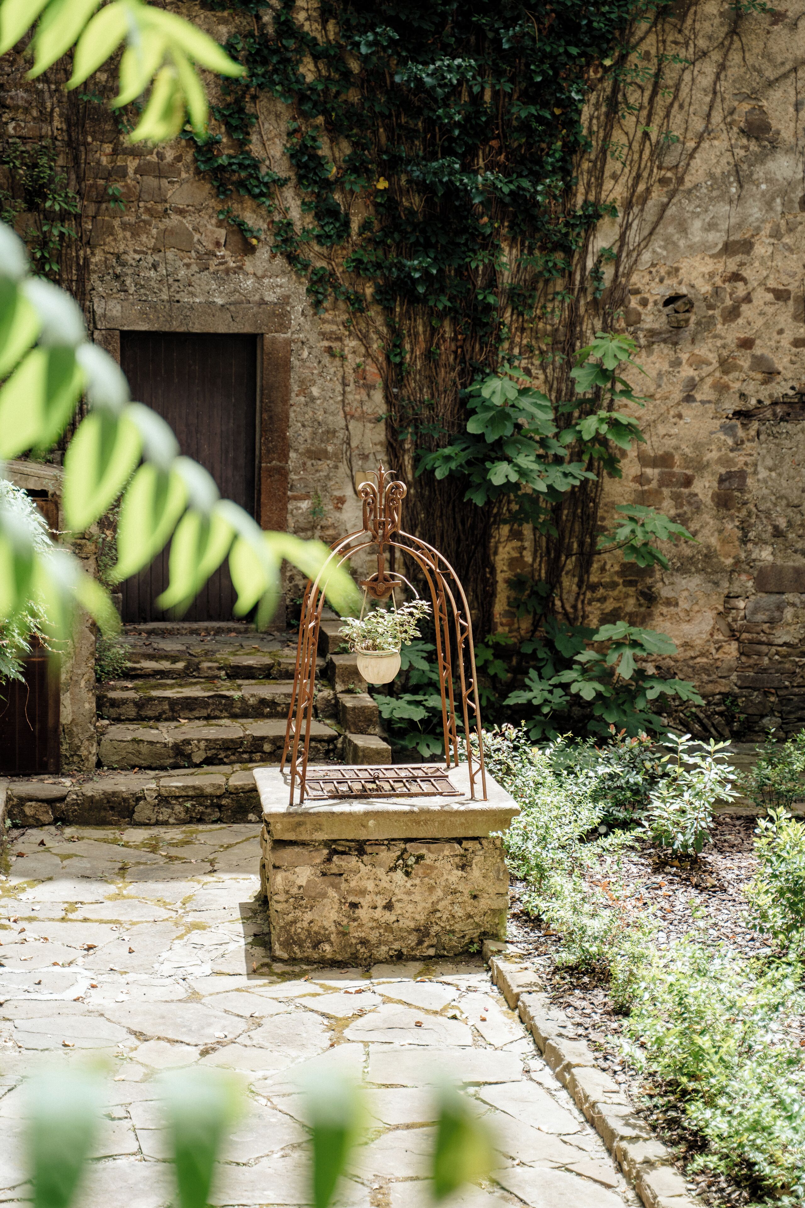 Un chemin de pierres menant à une porte rustique, entourée de verdure et surmonté d’une arche en métal ornée de jardinières suspendues.