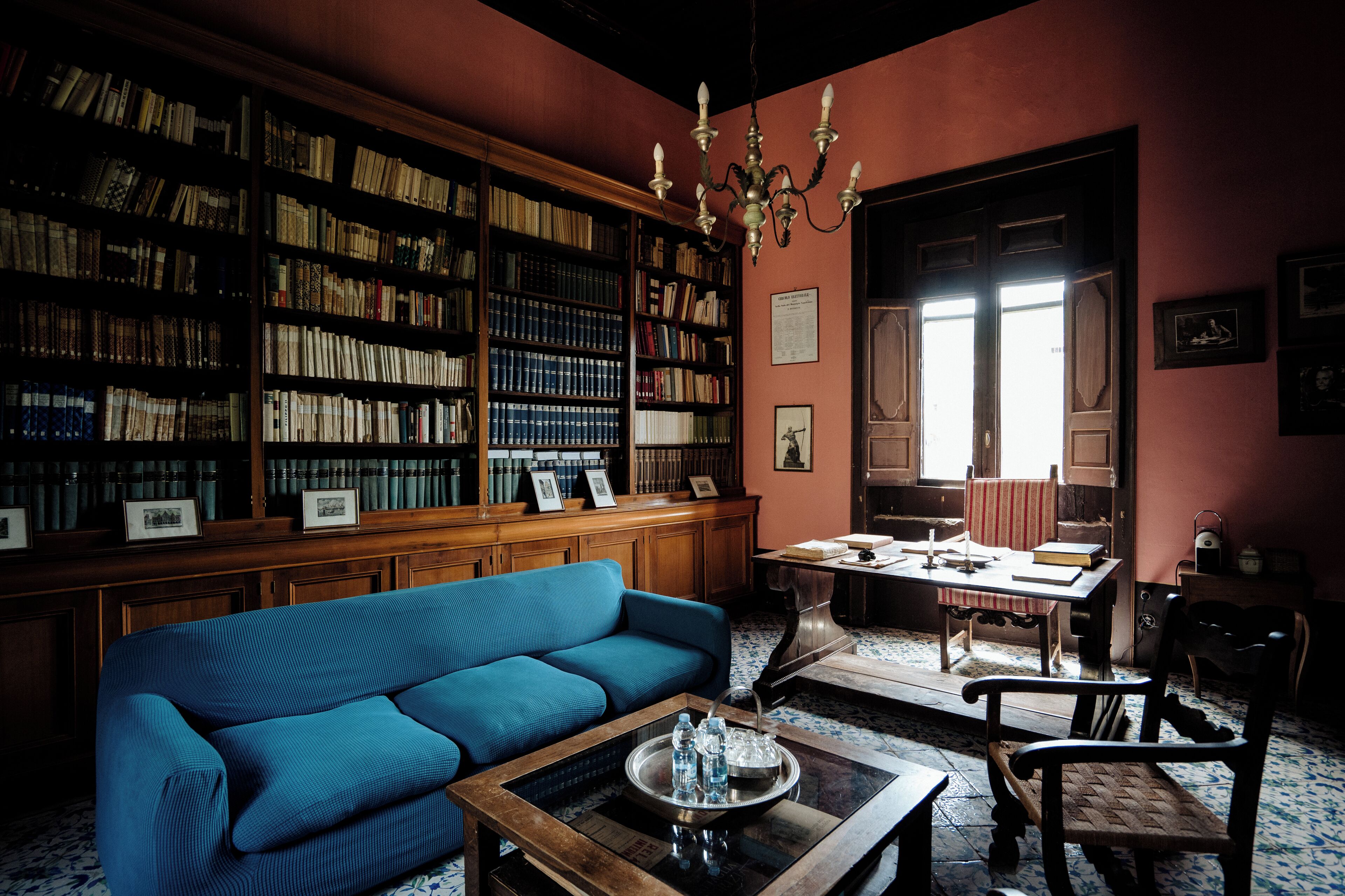 A library with bookshelves, a blue sofa, a wooden table, and a chandelier.