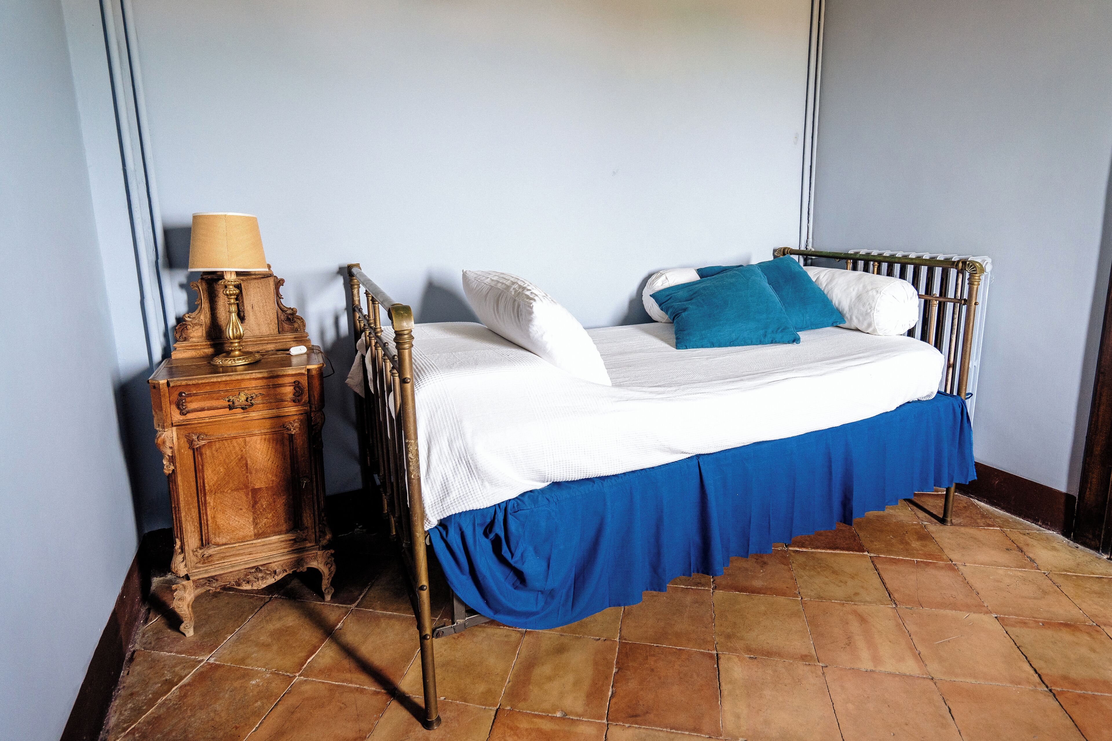 Una cama con colcha blanca y azul, una mesita de noche de madera con lámpara y un suelo de baldosas.