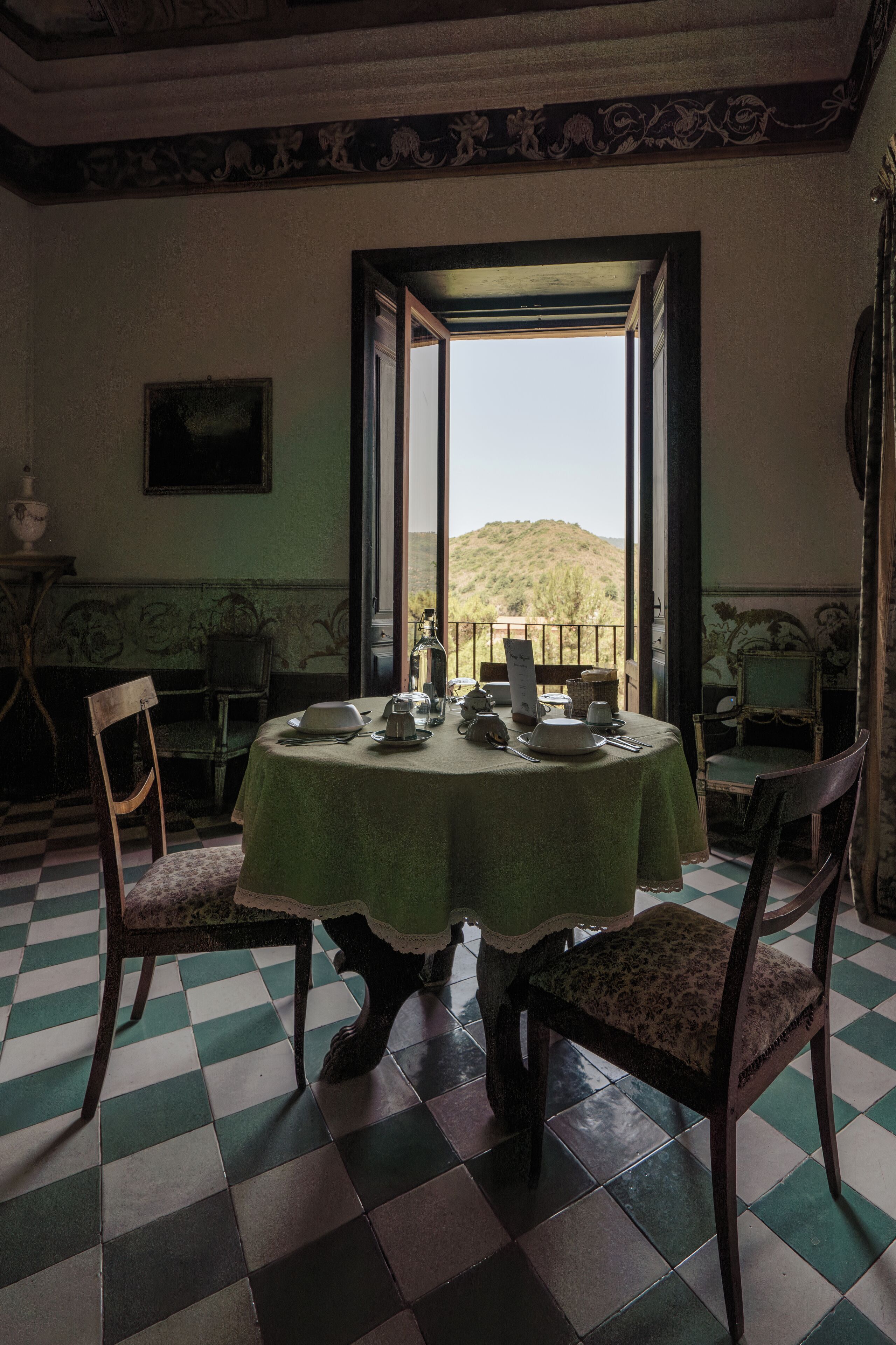 Comedor con una mesa preparada para cuatro personas, sillas, vista a una colina y piso de tablones de ajedrez.
