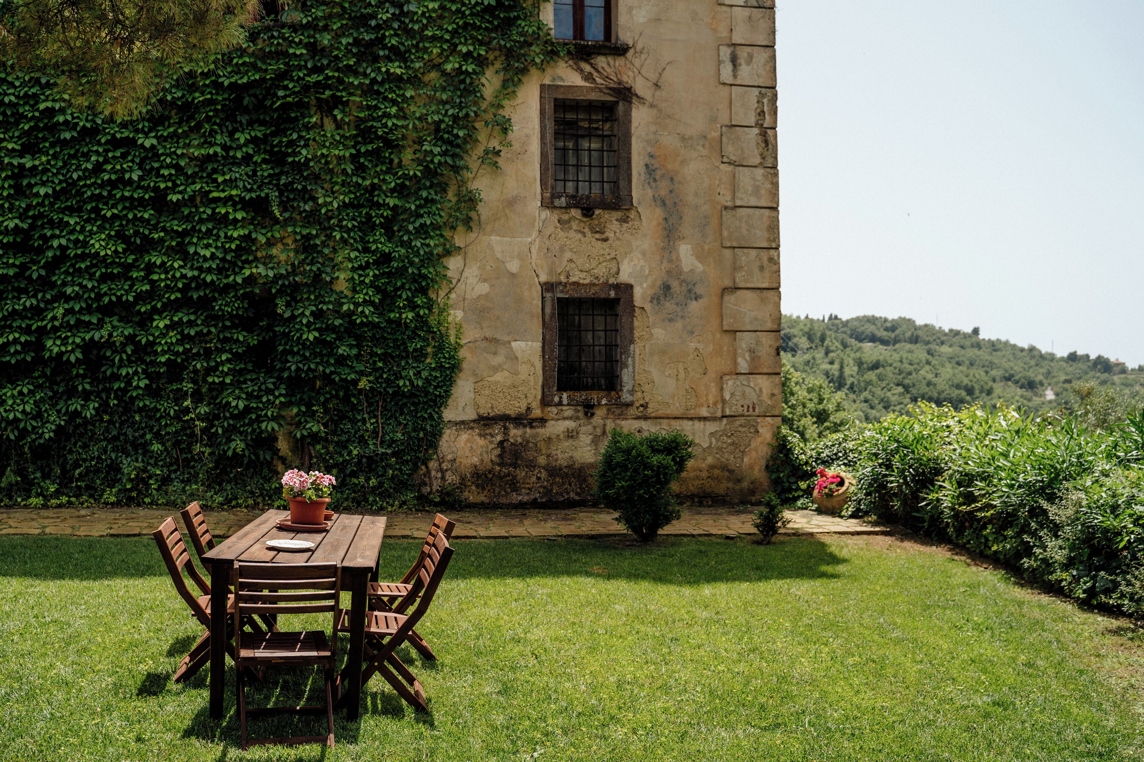 A rustic outdoor dining area with a wooden table and chairs, surrounded by lush greenery and a stone building with ivy.