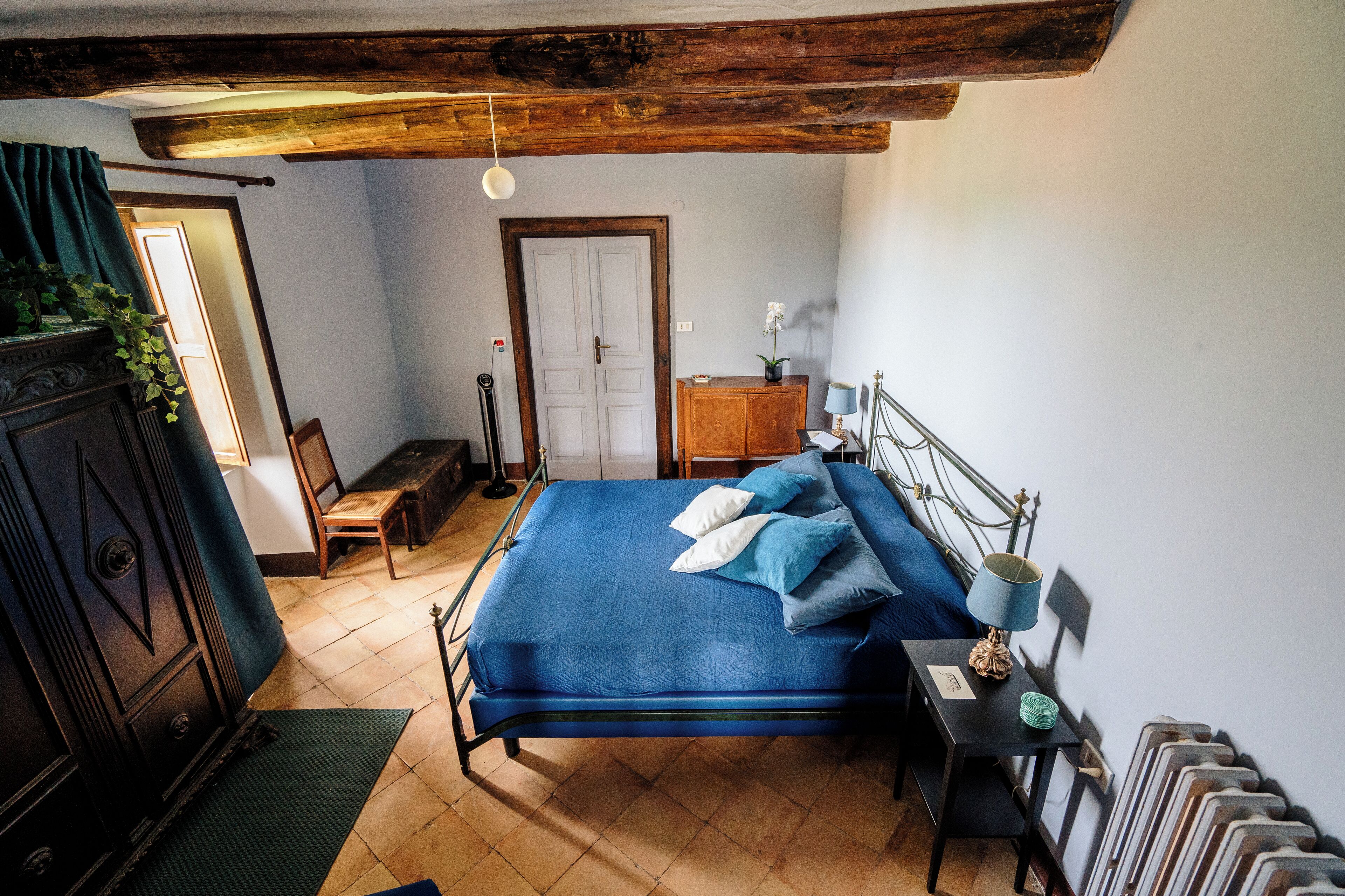 A bedroom with a wooden ceiling, a bed with blue bedding, a dresser, a radiator, and a door leading to another room.