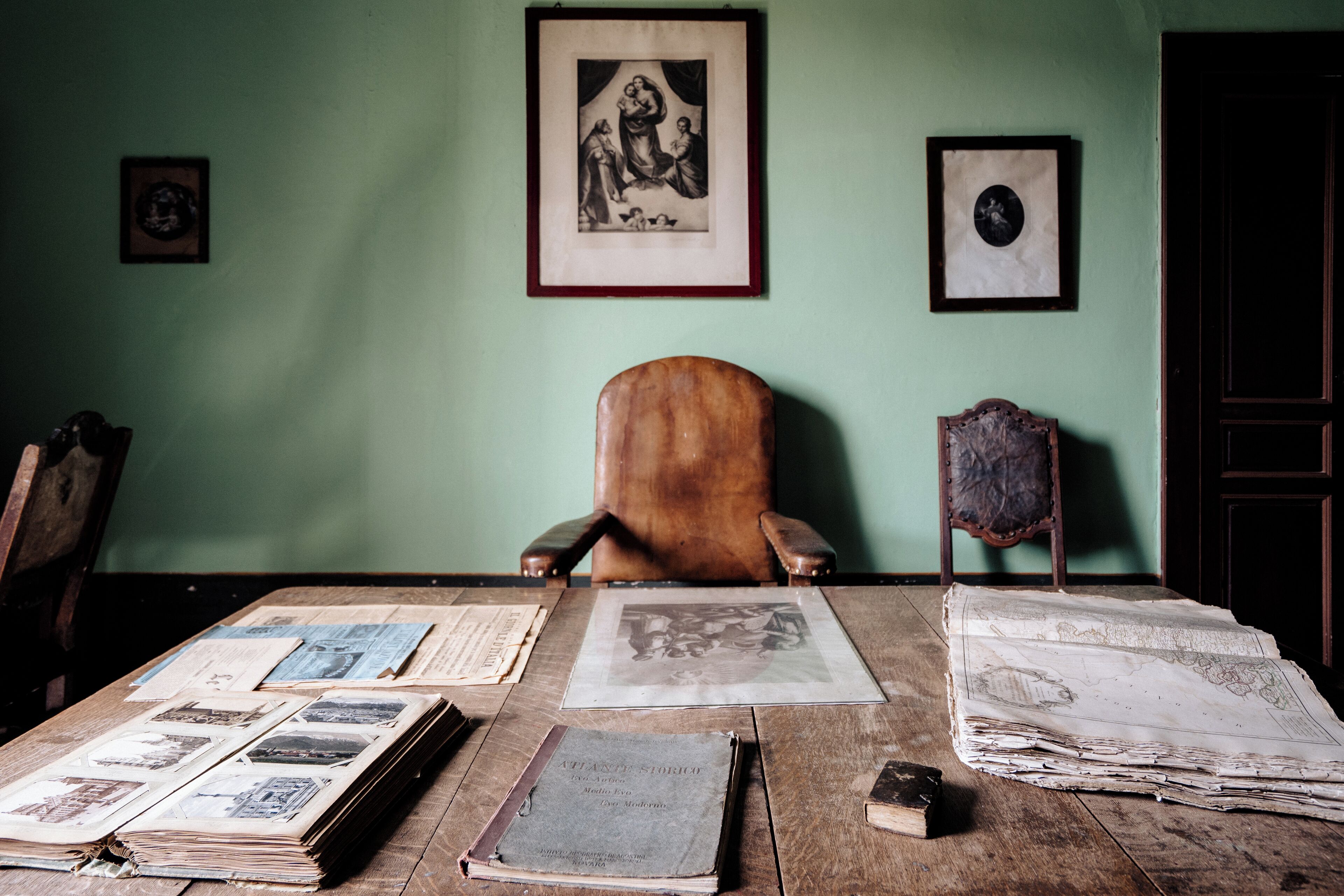 A room with a wooden desk, several old books, newspapers, and framed pictures on the wall.