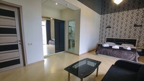 Una habitación de hotel moderna con una mesa de vidrio, un sofá, una cama con toallas ordenadas y un baño visible a través de una puerta abierta.