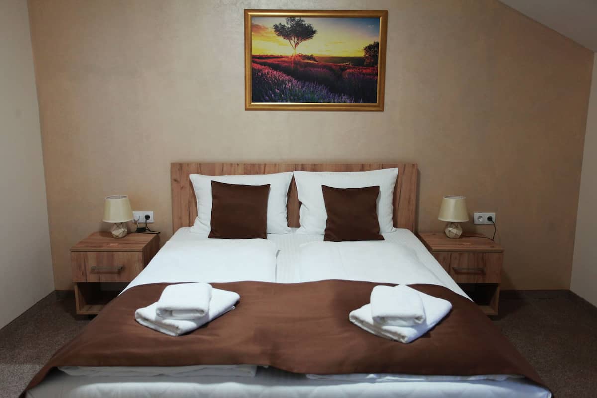 A neatly made bed with a brown bedspread and white linens, two brown pillows, and two white pillows. There are two bedside tables with lamps and a framed picture of a sunset on the wall above the bed.
