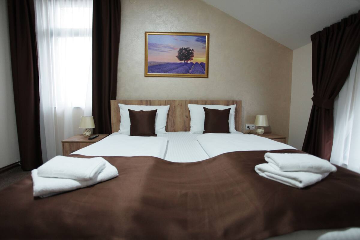 A double bed with a brown bedspread, two white pillows, and two brown pillows. There is a framed picture of a tree on the wall.